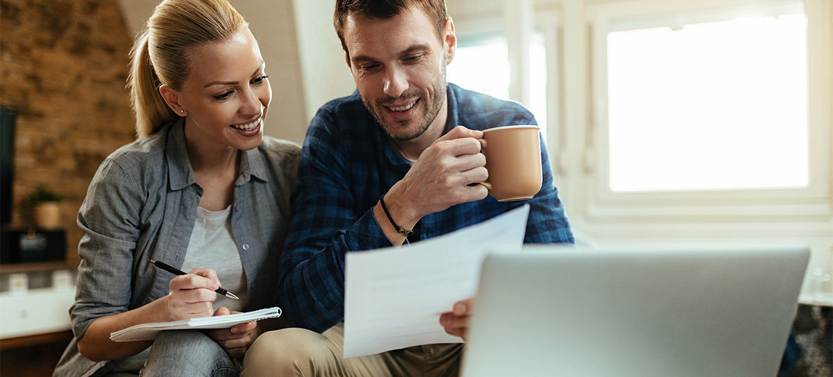 Couple smiling, looking at paper and taking notes with laptop in front of them in living room.